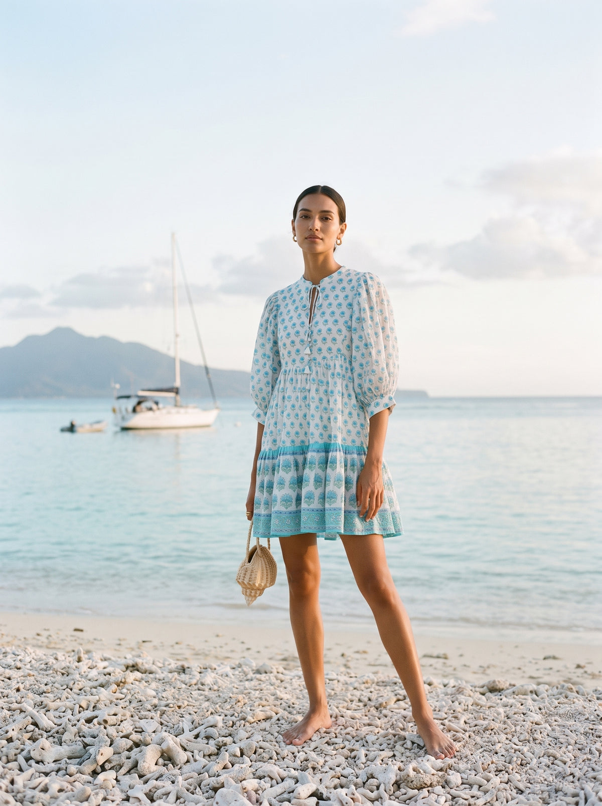 A woman stands barefoot on a pebble beach wearing a light blue and white patterned dress, holding a seashell, with a sailboat and ocean in the background.