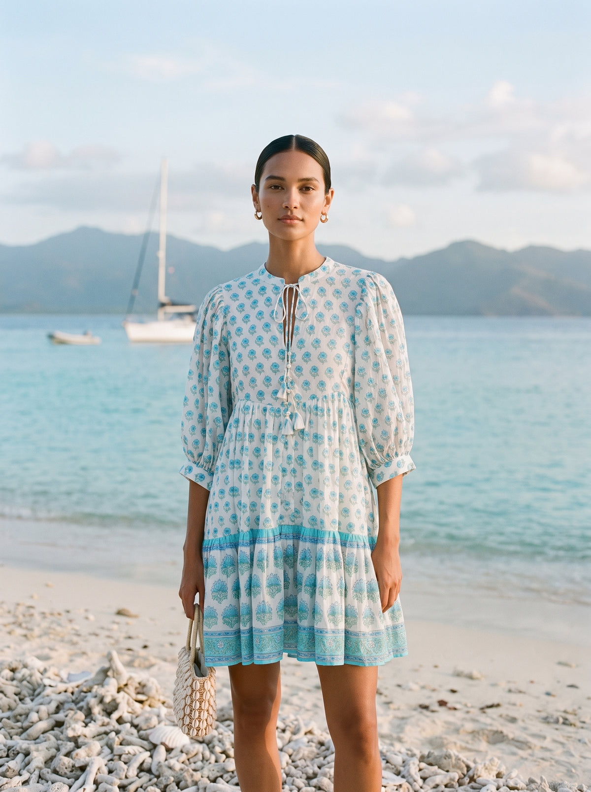 A woman stands on a beach wearing a light blue and white patterned dress with puff sleeves and a tie neckline. The background shows turquoise water, distant mountains, and a sailboat.